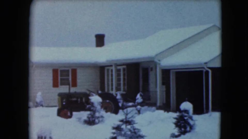 1961 Jamestown, Wisconsin frame showing the farmhouse front, red shutters, garage, snow-covered yard, shrubs, and vintage tractor.