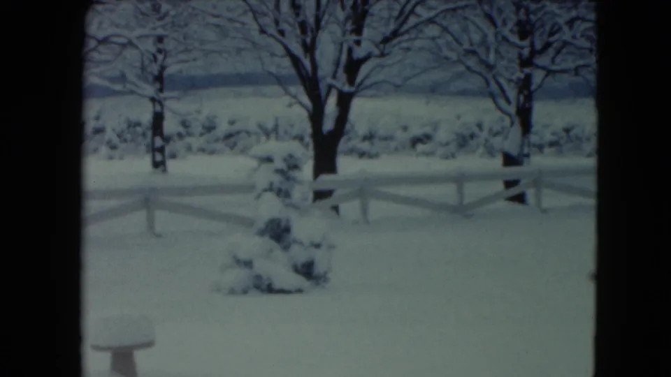 1961 Jamestown, Wisconsin 8mm frame showing a snowy yard, white fence, bare trees, and an evergreen shrub in a rural winter setting.