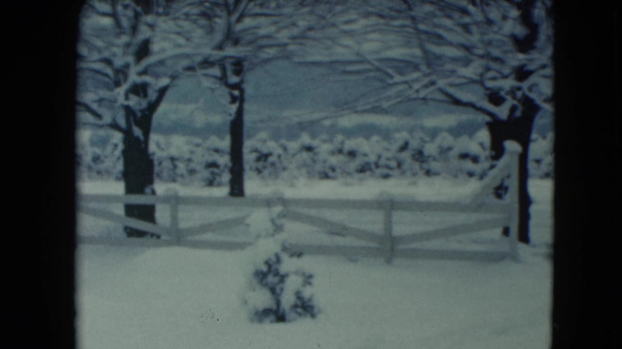 1961 Jamestown, Wisconsin 8mm frame showing a snowy yard, white fence, bare trees, low shrubs, and distant hills under a muted sky.