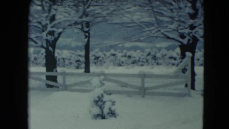 1961 Jamestown, Wisconsin 8mm frame showing a snowy yard, white fence, bare trees, low shrubs, and distant hills under a muted sky.