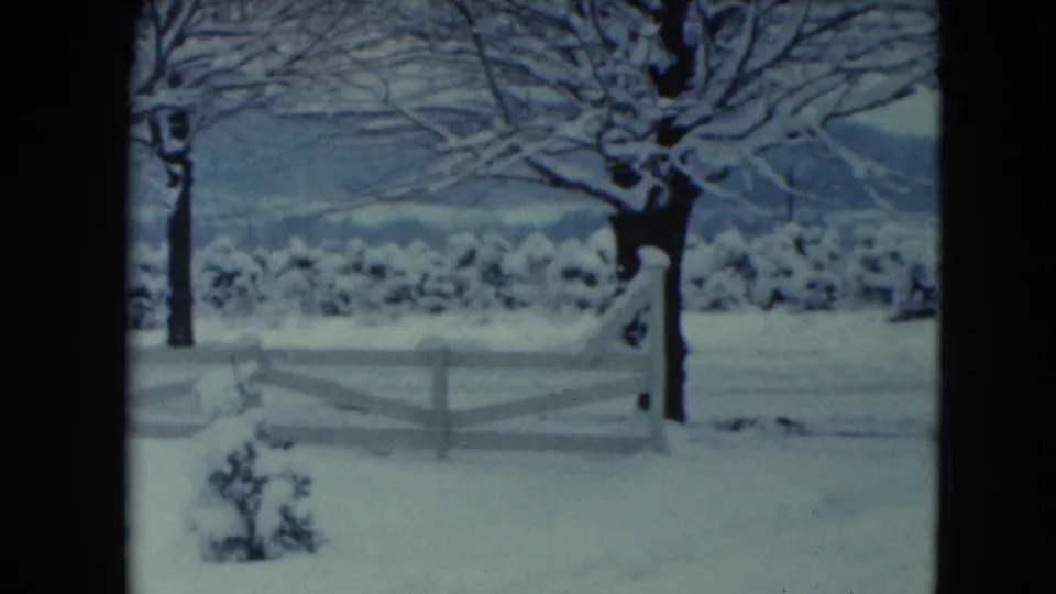 1961 Jamestown, Wisconsin home-movie frame with a white fence crossing a snow-covered yard beneath bare winter trees.
