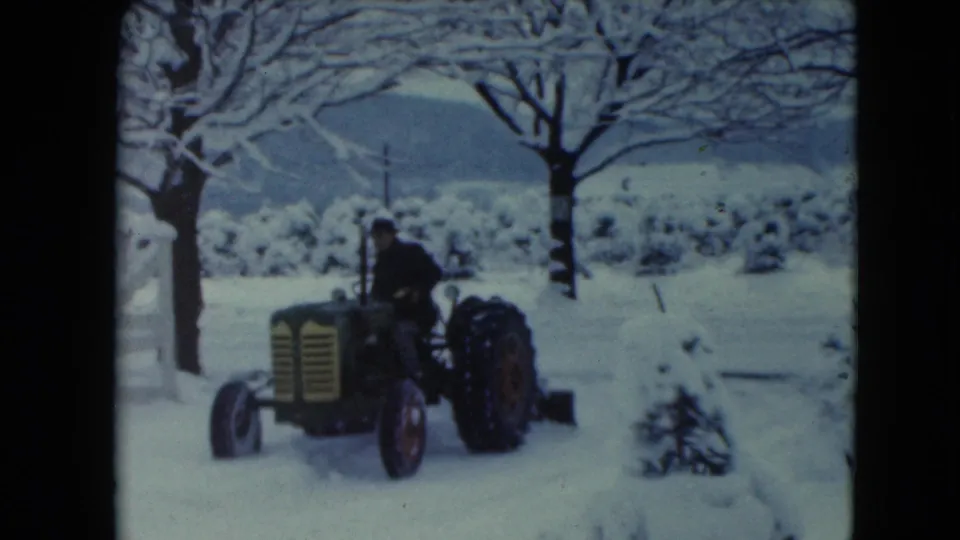 A man in dark winter clothing drives the tractor and blade past a snowy tree in the 1961 rural Jamestown scene.