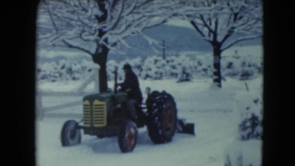 A side view shows the man operating the green tractor and blade through the snowy Wisconsin yard.