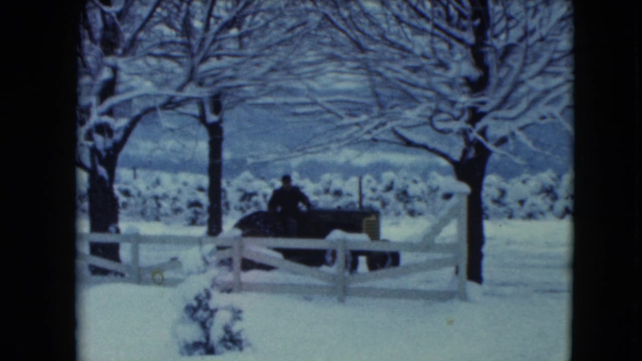 1961 Jamestown, Wisconsin home-movie frame showing a person on a tractor near a white fence in a snow-covered rural field.