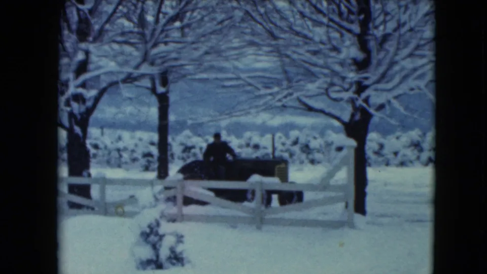1961 Jamestown, Wisconsin home-movie frame showing a person on a tractor near a white fence in a snow-covered rural field.