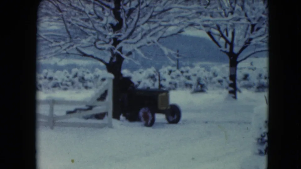 A person drives a vintage tractor pulling a plow past a white fence in the 1961 snowy Wisconsin farm scene.