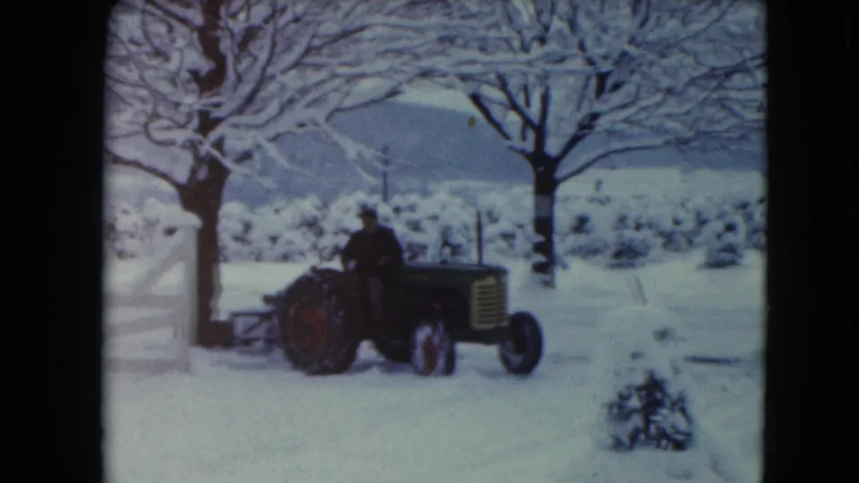 The driver continues across the snowy Jamestown field on a tractor pulling a plow, framed by trees and a white fence.