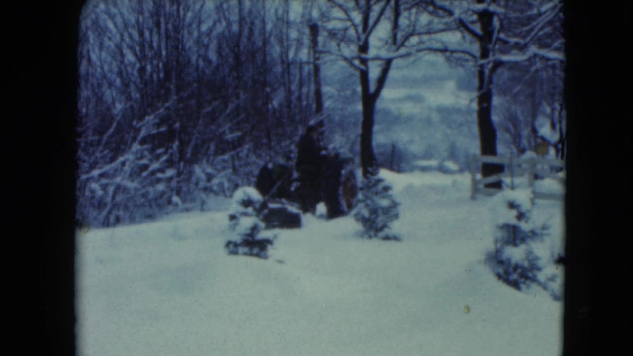 Dark-clothed person on a tractor in a snowy rural yard with trees and a white fence.