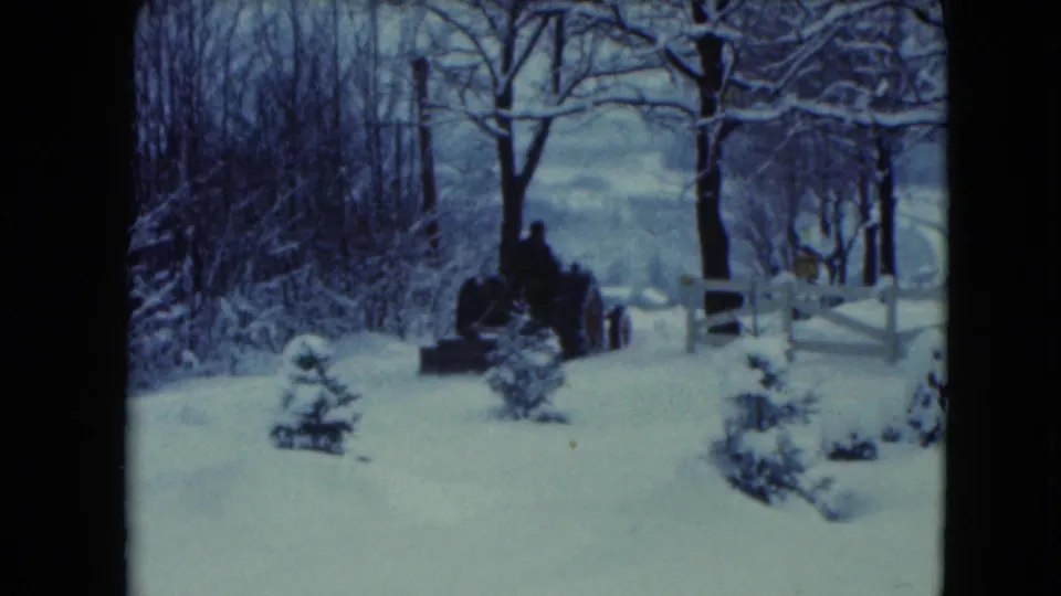 Closer 8mm view of the tractor and driver crossing the snow-covered rural property in Jamestown, Wisconsin.