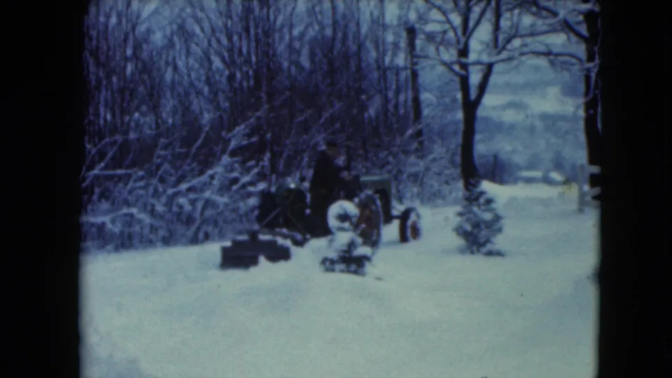 The 8mm frame shows a man maneuvering a tractor through a snowy rural yard in Jamestown, Wisconsin.