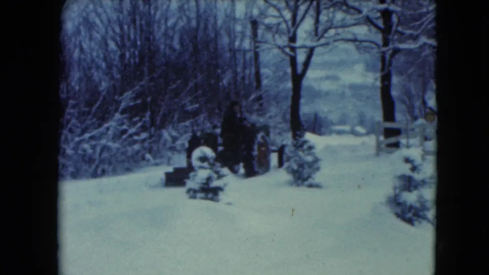 A man on a tractor appears farther across the snowy yard, with bare trees and a low fence visible nearby.