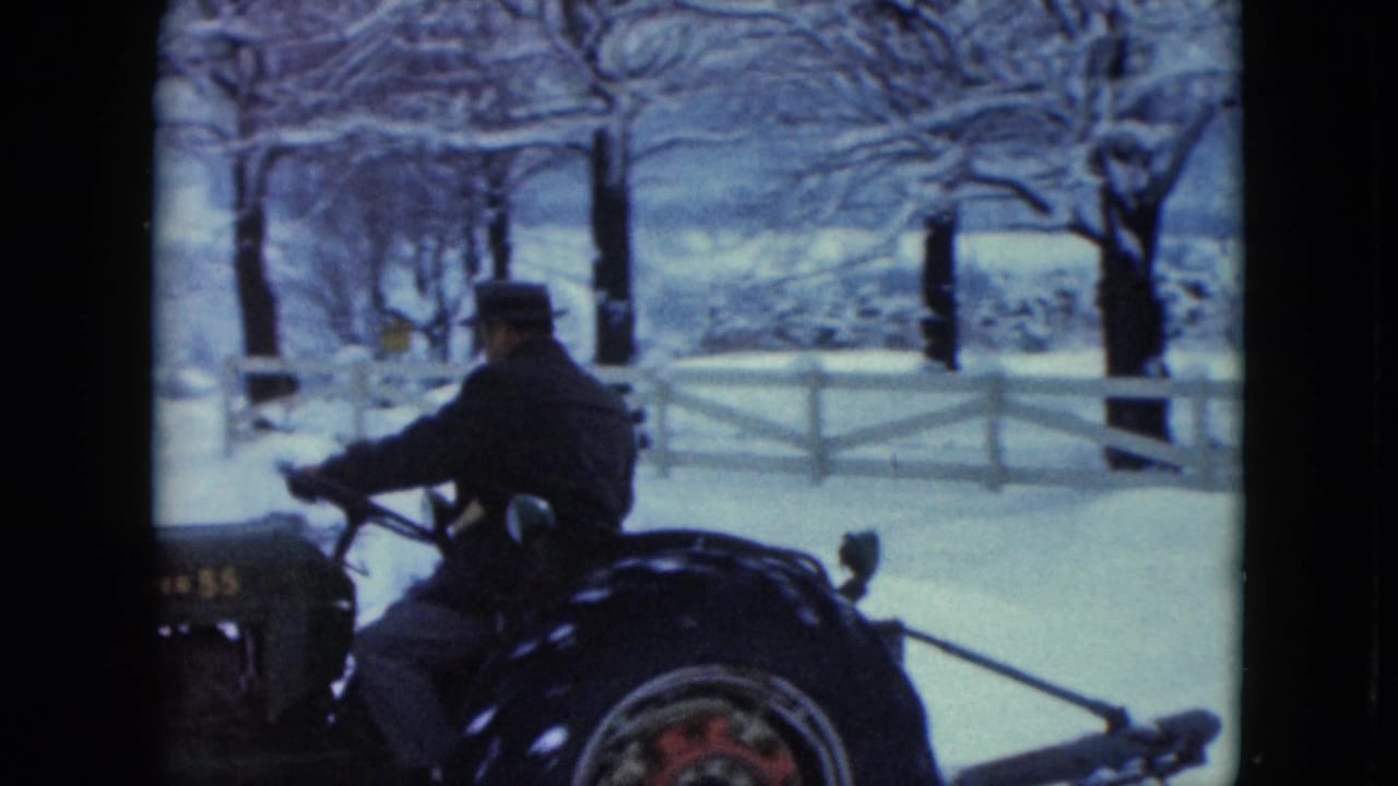 In Jamestown, Wisconsin, 1961, a man in a dark coat and hat drives a tractor through a snowy rural yard near a white fence.