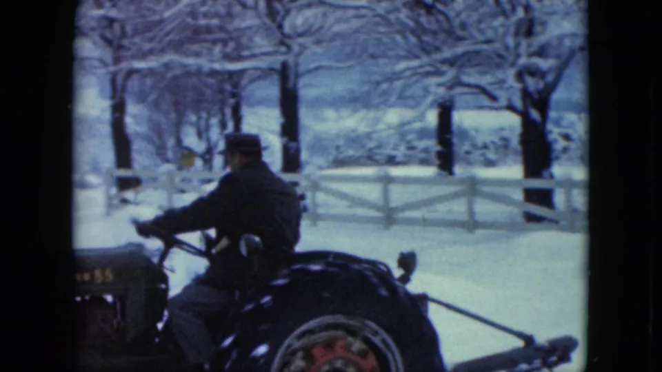 In Jamestown, Wisconsin, 1961, a man in a dark coat and hat drives a tractor through a snowy rural yard near a white fence.