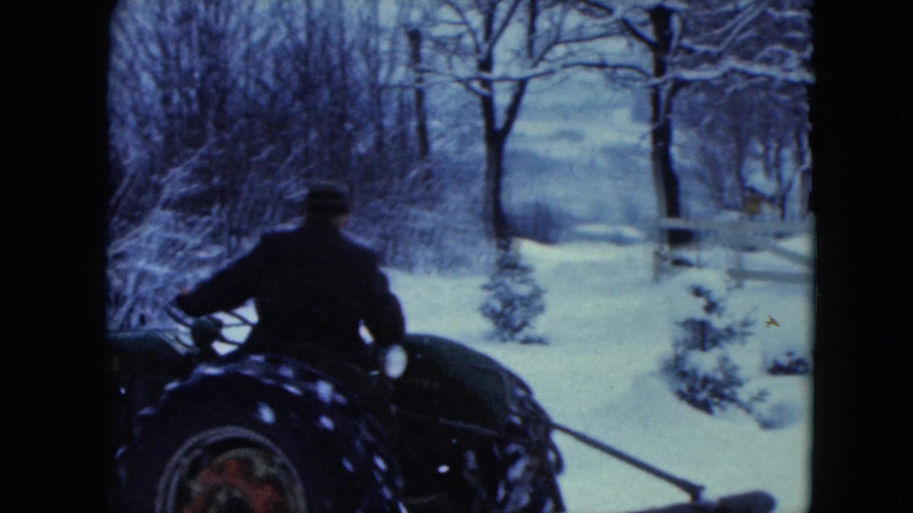 Rear view of a man on a tractor moving across a snowy yard with trees behind him.