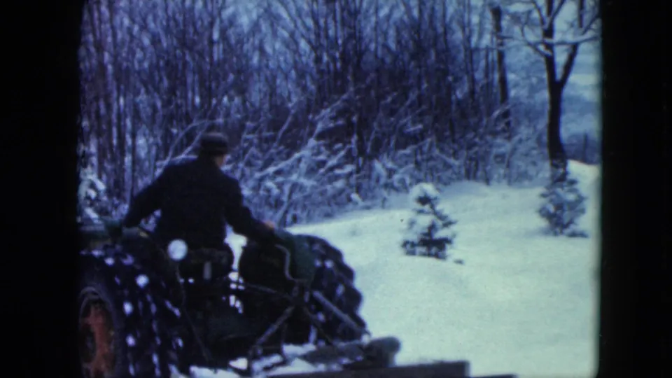 A rear view shows the man steering the tractor across snow near winter brush and small evergreens in Jamestown, Wisconsin.