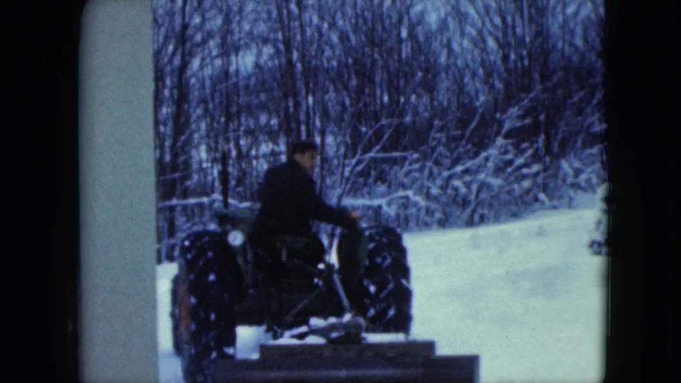 The man looks partly toward the camera while operating the tractor near a building edge in the snowy 1961 Wisconsin scene.