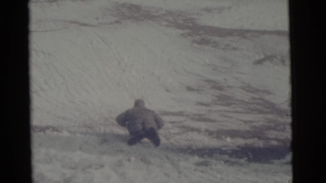 In the supplied 1961 Camden, New Jersey home-movie frame, a bundled person begins sledding away across a snowy slope.