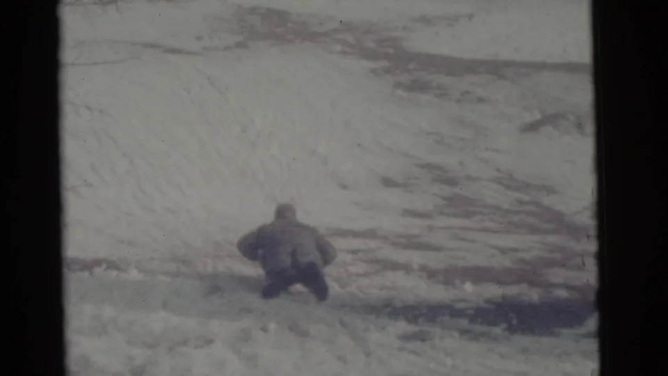 In the supplied 1961 Camden, New Jersey home-movie frame, a bundled person begins sledding away across a snowy slope.