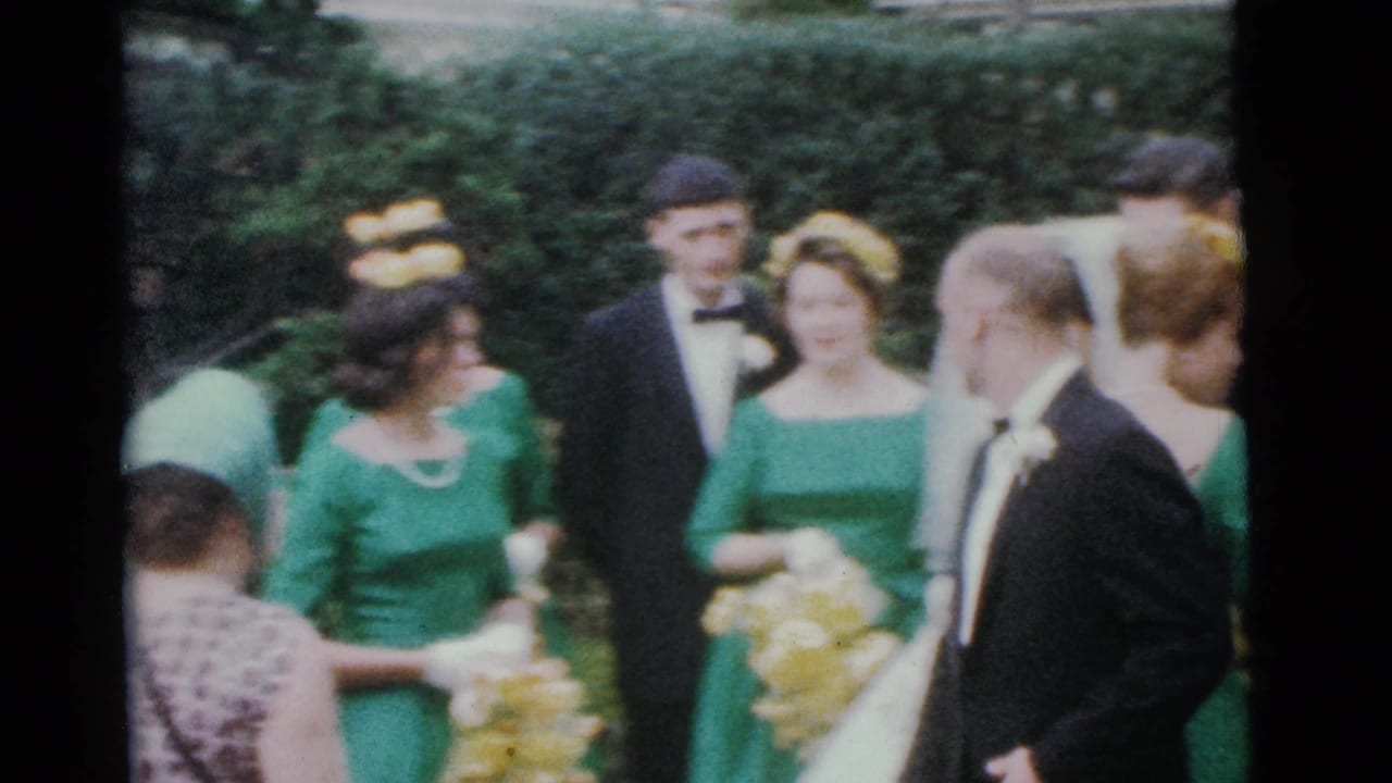 Wedding attendants in green dresses and tuxedos stand near hedges with yellow flowers.