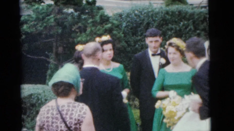 1961 Camden wedding scene with the bride partly visible among bridesmaids in green dresses, tuxedoed men, and guests near hedges.