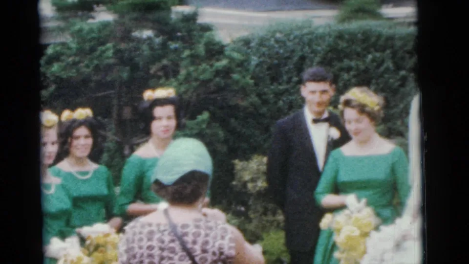 1961 Camden wedding party frame showing the bride in a veil, groom in tuxedo, bridesmaid in green, and best man beside hedges.