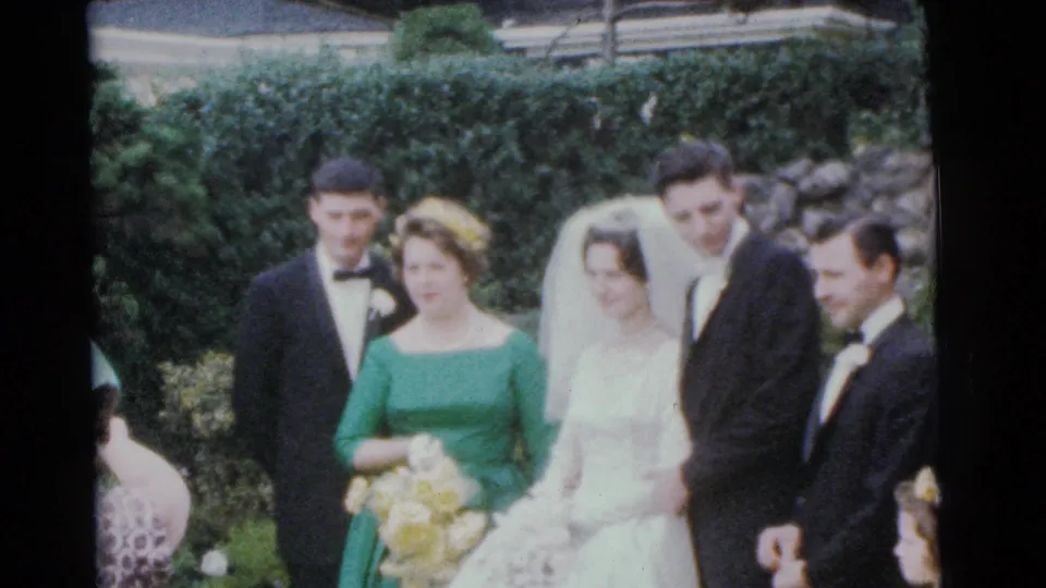 1961 Camden home-movie frame of the bride and groom posed with a bridesmaid in green, best man, and another suited attendant outdoors.