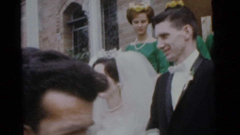 The groom smiles beside the veiled bride during a 1961 Camden church wedding scene, with a bridesmaid visible in the background.