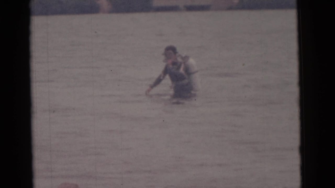 1961 Camden, New Jersey home-movie frame showing a male scuba diver wading in shallow water with a light tank.