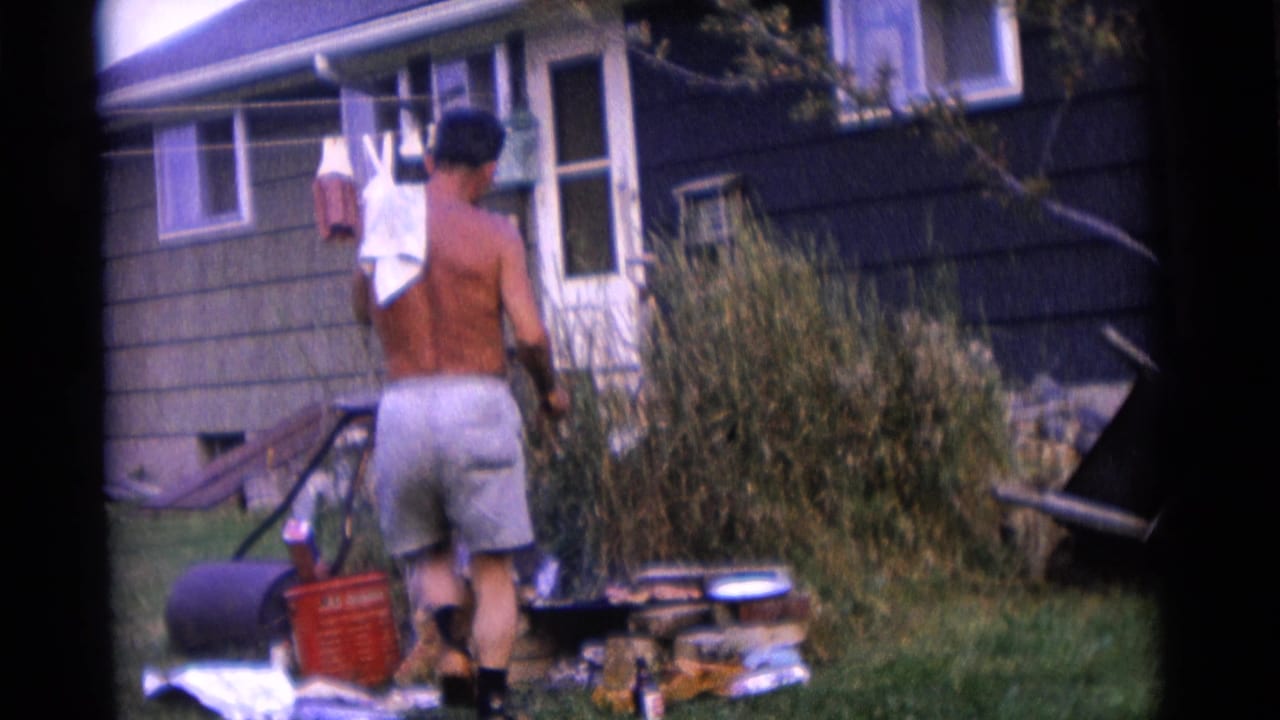 Shirtless man in light shorts stands by a backyard grill near a dark-sided house.