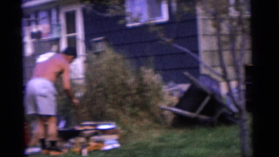 1961 Camden backyard frame with a man leaning toward the grill near shrubs, lawn, and house windows.