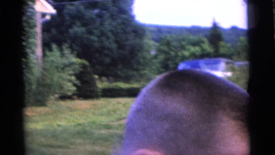 1961 Camden home-movie frame showing a backyard lawn, trees, distant yard features, and a close partial head in the foreground.