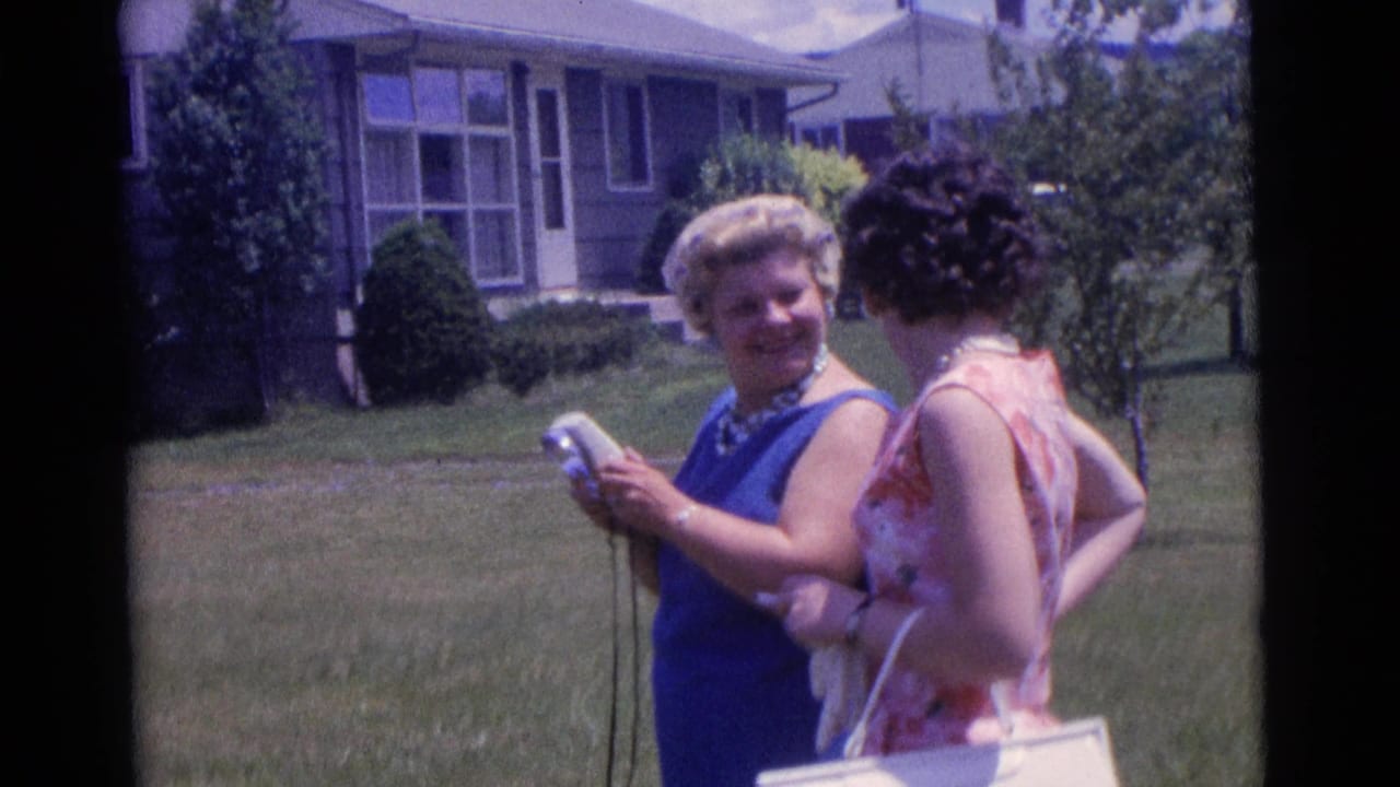 In Camden, New Jersey in 1961, two women stand in a suburban yard while one smiles and holds a small camera.