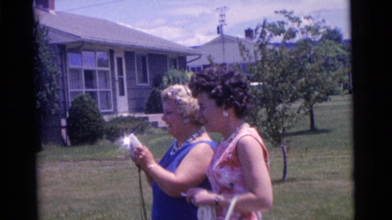 A woman in blue looks down at a small camera beside another woman on a lawn.