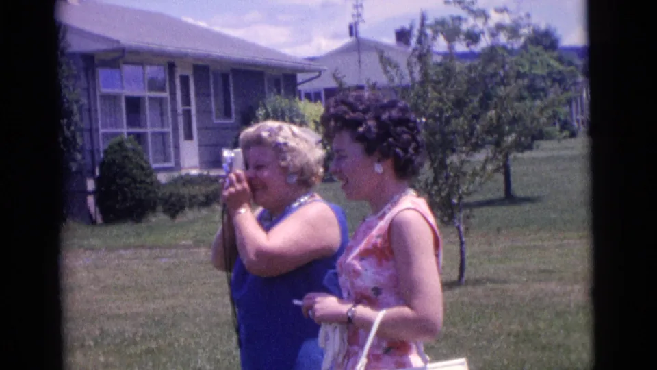 In Camden, New Jersey in 1961, one woman raises a small camera to eye level as another woman stands beside her on the lawn.