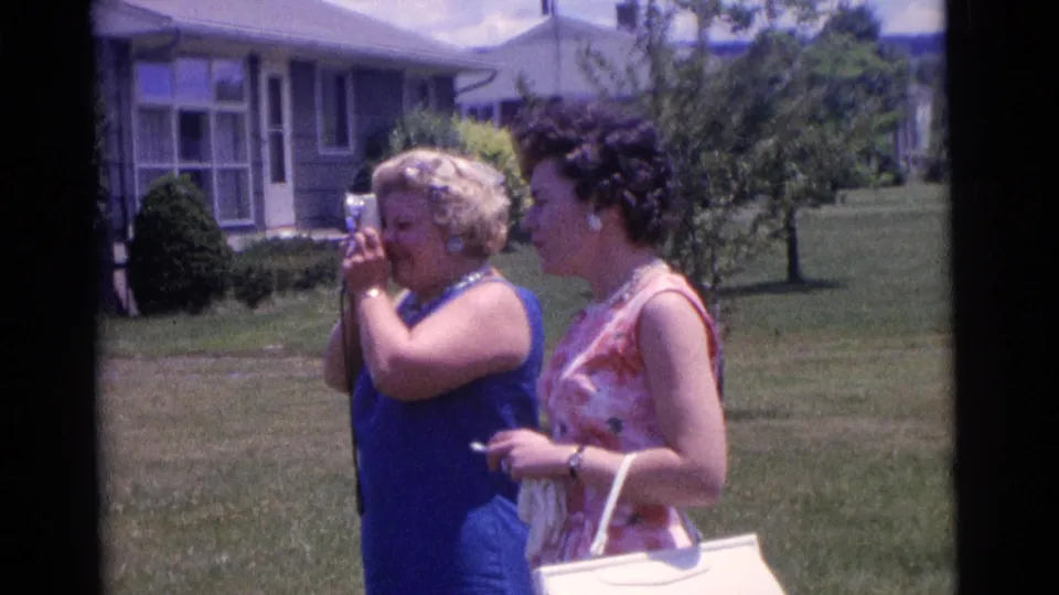 The 1961 Camden home-movie frame shows one woman aiming a small camera while her companion stands beside her in a suburban yard.
