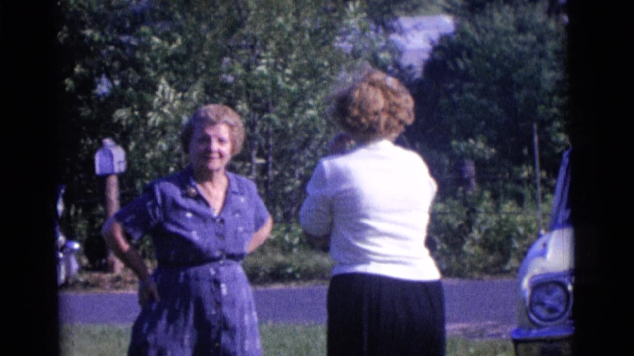 Two women face each other beside a road with a car at the edge of frame.