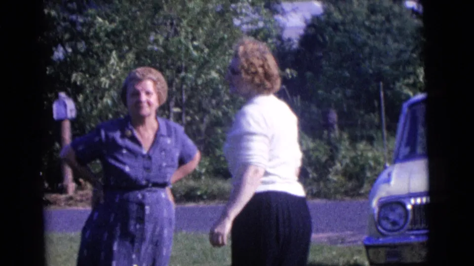 The women continue their outdoor conversation in 1961 Camden, with trees, a mailbox, lawn, road, and car framing the scene.