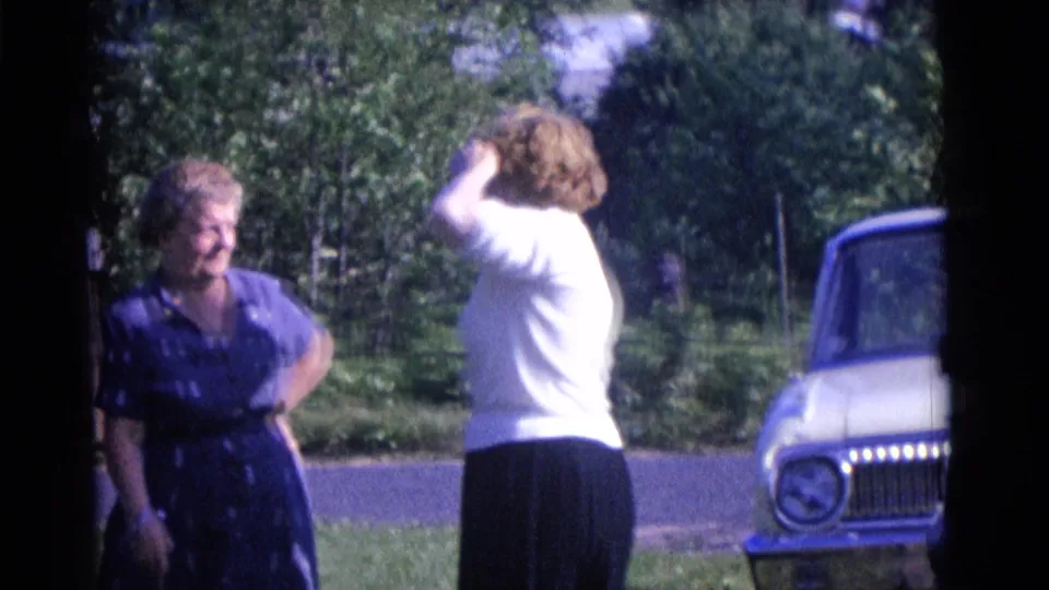 A woman in a white blouse raises her arm to adjust her hat or hair as the other woman stands beside her in the 1961 Camden scene.