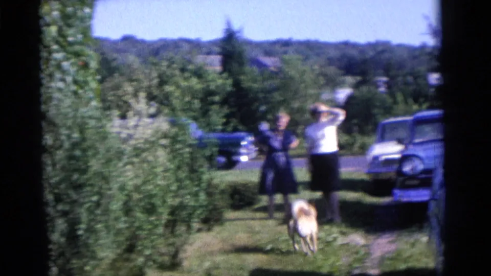 1961 Camden home-movie frame of two adults in a yard, a dog on the grass, trees, shrubs, and mid-century cars nearby.