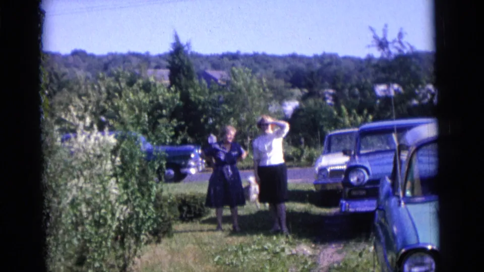 1961 Camden yard scene with two adults, a dog, leafy shrubs, a blue car at the edge, and other parked cars beyond.
