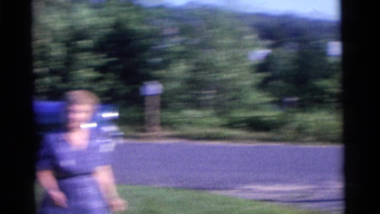 Blurred woman moves across a lawn with a blue car and shrubs behind her.