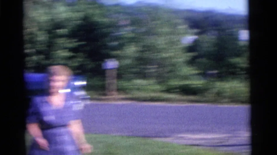 A handheld 8mm frame shows a woman in a blue dress in motion near a blue car and green shrubs in Camden, New Jersey.