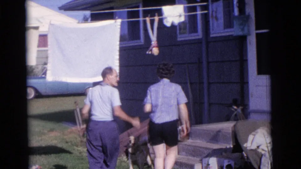 Two adults move through a Camden backyard with laundry hanging overhead, a house exterior behind them, and a dog near the grass.