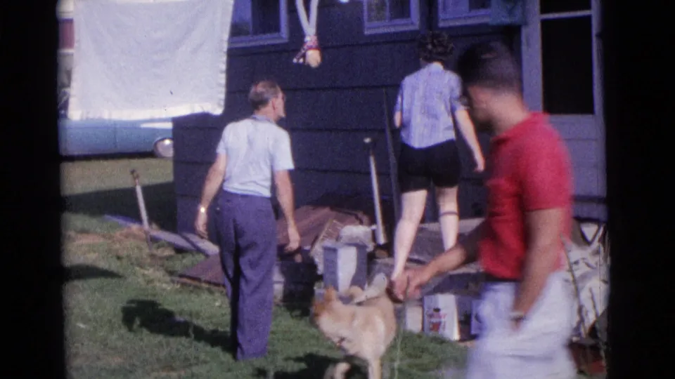 A 1961 Camden backyard frame shows adults near a dog and clothesline while a young man in a red shirt passes in the foreground.