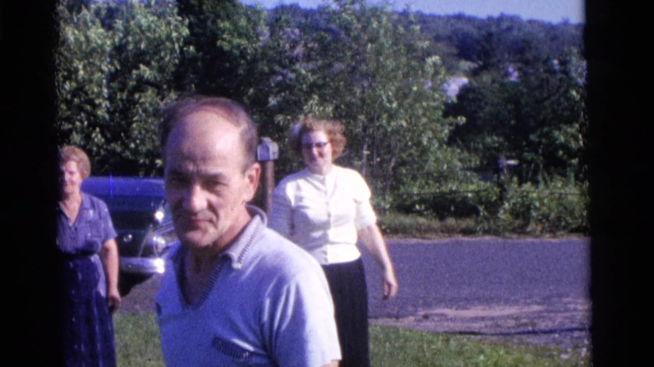 Man in a light shirt stands near the camera as two women walk behind him on a lawn.