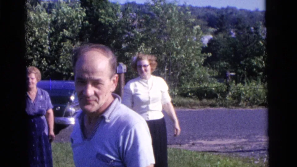 In Camden, New Jersey, 1961, a man in a light shirt stands in front while two women approach from a lawn near a blue car.
