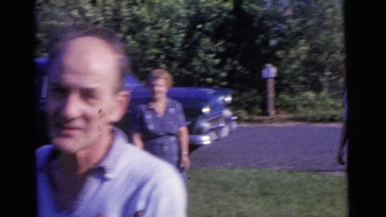 Man passes close to the camera with two women and a blue car behind him.