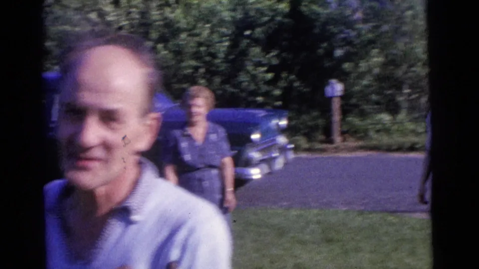 The man moves close to the lens while two women remain behind him near a parked blue automobile in the 1961 Camden home-movie scene.