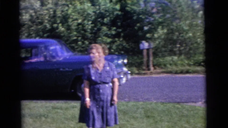 A woman in a blue dress walks toward the camera on a lawn, with a blue period car, road, trees, and mailbox behind her.