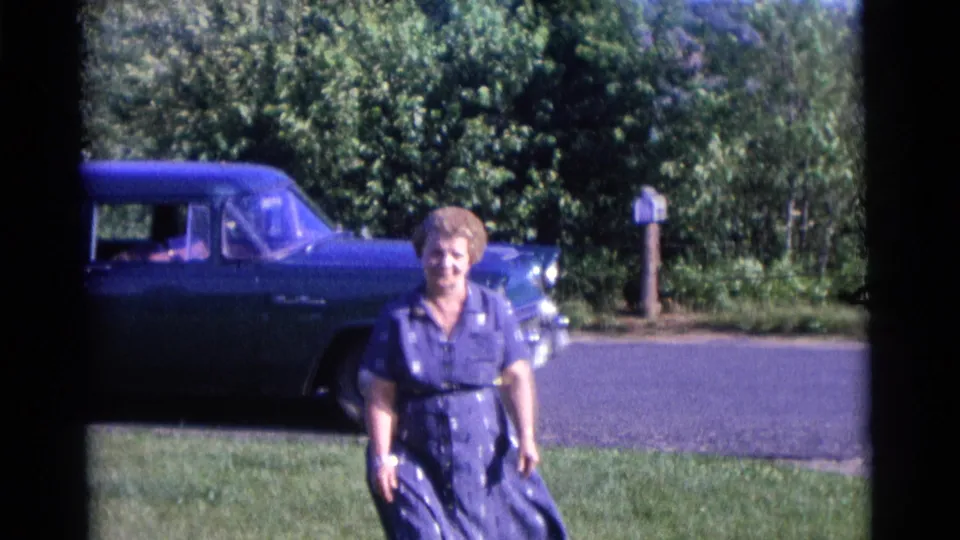 In the Camden 1961 footage, a woman in a blue dress smiles on the lawn with a blue automobile, trees, road, and mailbox in the background.